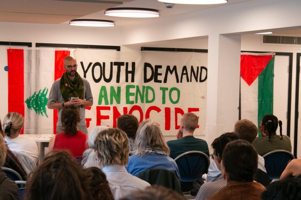 A man speaks to a seated crowd. The banner behind him reads: "Youth demand an end to genocide."