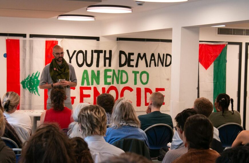 A man speaks to a seated crowd. The banner behind him reads: "Youth demand an end to genocide."