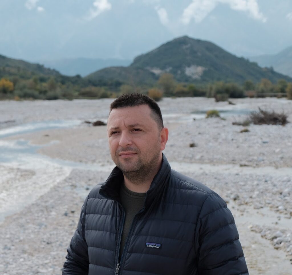 Man stands in front of a river, with a hill in the background.