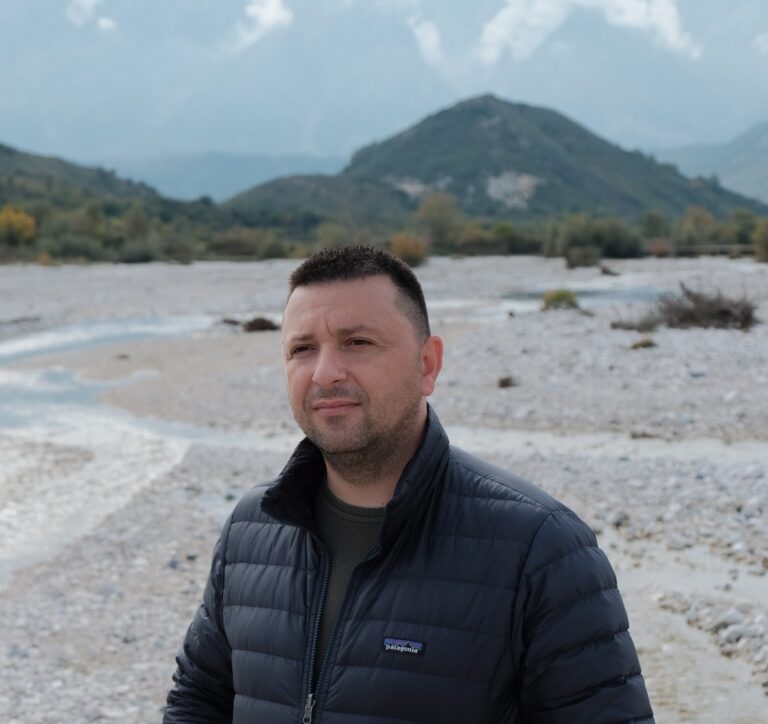 Man stands in front of a river, with a hill in the background.
