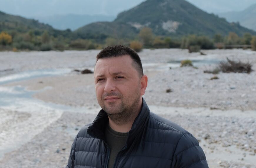 Man stands in front of a river, with a hill in the background.