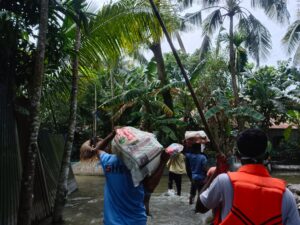 A small group of young volunteers, one of them wearing a t-shirt with SHBO's logo on the back, carry bags of supplies over their shoulders and heads through a waterlogged village with lush vegetation.