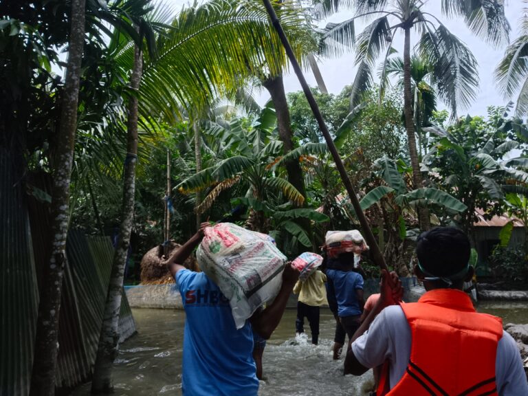 A small group of young volunteers, one of them wearing a t-shirt with SHBO's logo on the back, carry bags of supplies over their shoulders and heads through a waterlogged village with lush vegetation.