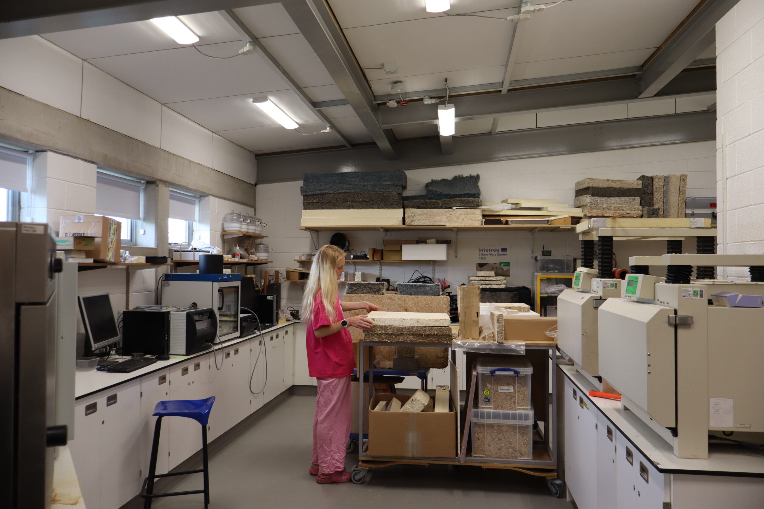 Joni Wildman, a young woman with a pink t-shirt and trousers, handles plaques of mycelium-based materials in a lab.