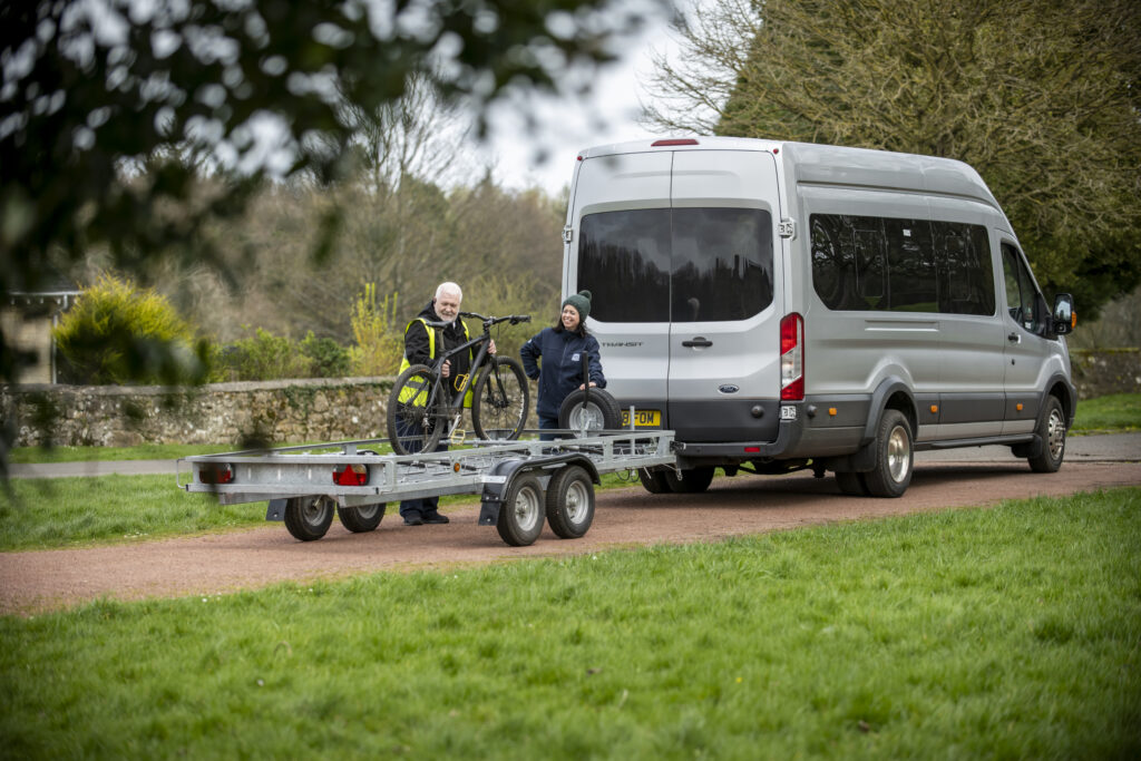 A bicycle is loaded onto a trailer behind a silver minibus