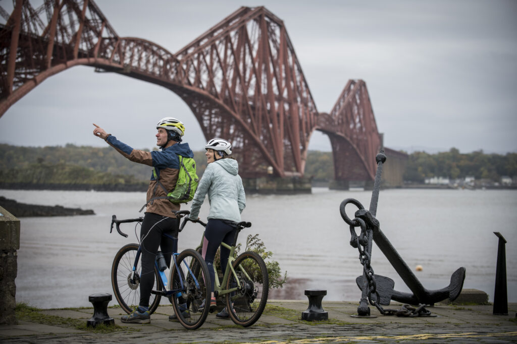 two cyclists pictured in the foreground,  with a large red bridge in the background
