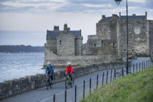 Two cyclists on a road in front of an old, stone building and a body of water