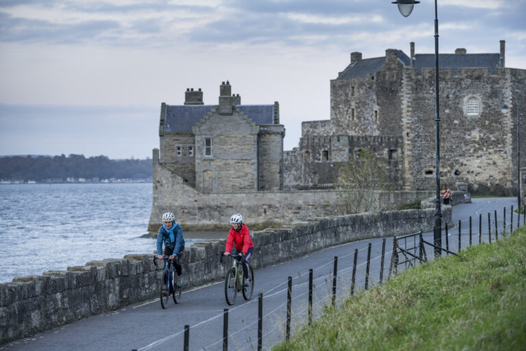 Two cyclists on a road in front of an old, stone building and a body of water