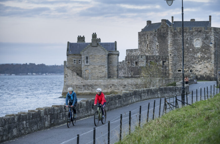 Two cyclists on a road in front of an old, stone building and a body of water