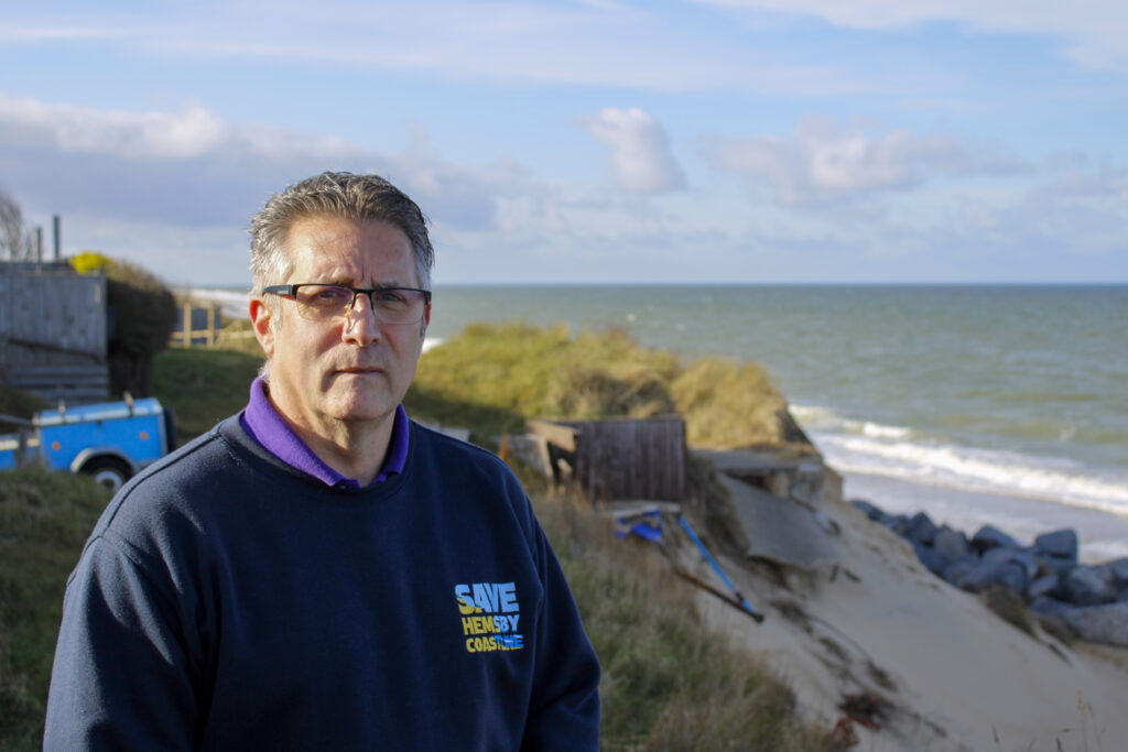 Man in a blue jumper stands in front of a crumbling coastline, with the sea behind him