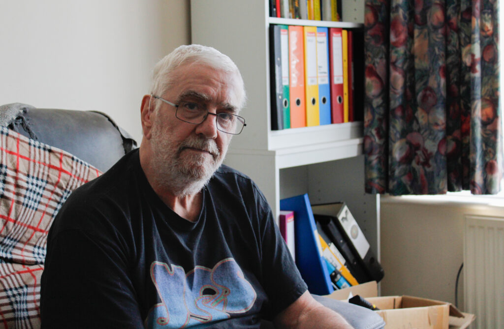 Man in a black 'Yes' t-shirt sits on a black sofa with a shelf behind him