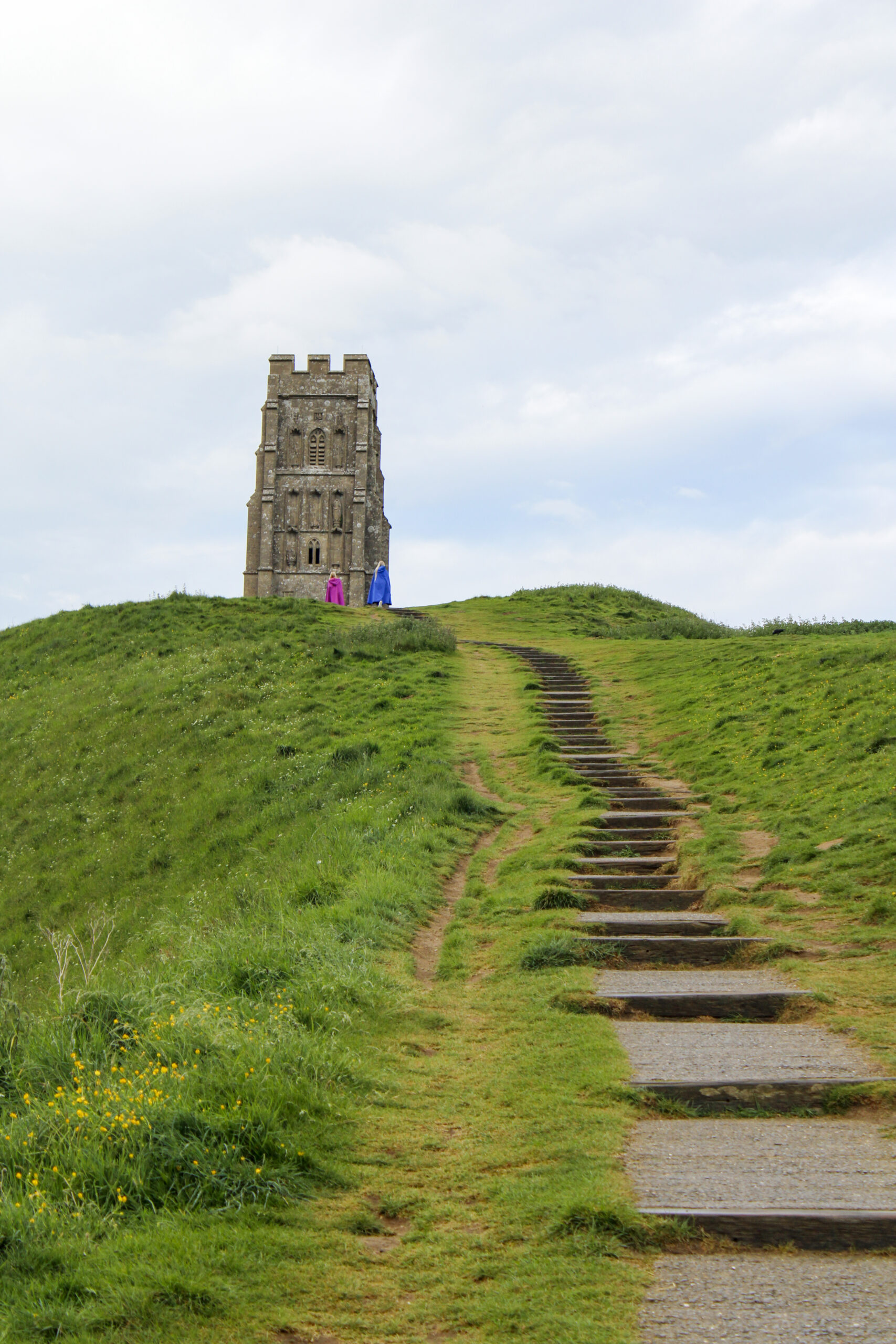 A castle turret on top of a green hill. Two people in pink and yellow robes stand under the turret.