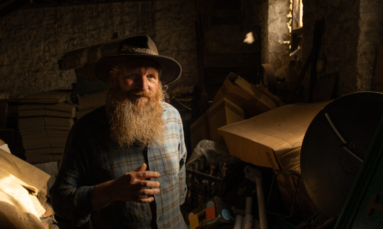 Bearded man wearing a wide-brimmed hat, stands in warm half-light inside a workshop