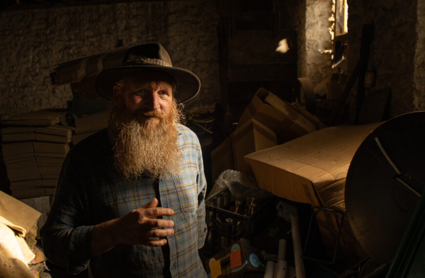 Bearded man wearing a wide-brimmed hat, stands in warm half-light inside a workshop