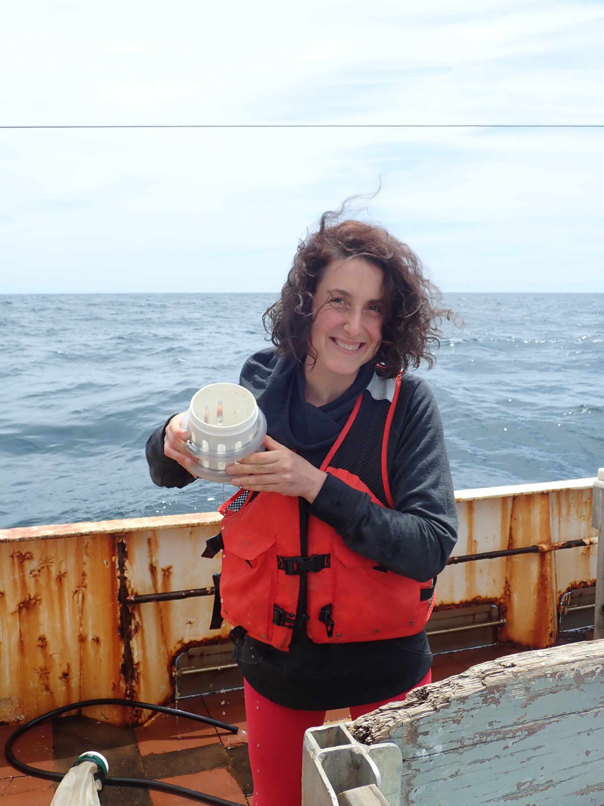 A woman holding a container filled with plankton. She wears a red fishing suit, and stands on board of a ship, with sea in the background