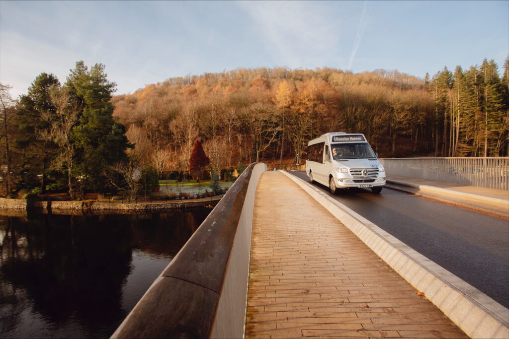 A silver bus crosses a bridge with a forest in the background