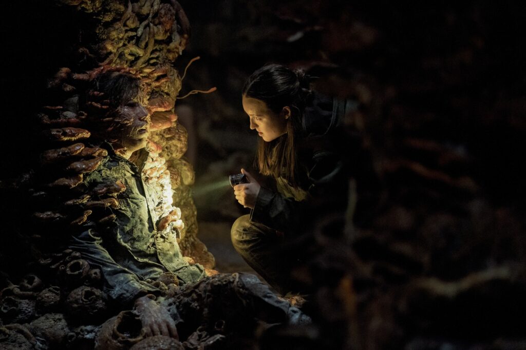 A young woman (Ellie, played by Bella Ramsey) points a flashlight at a cordyceps-infected man, who is infused with a wall covered in the fungus