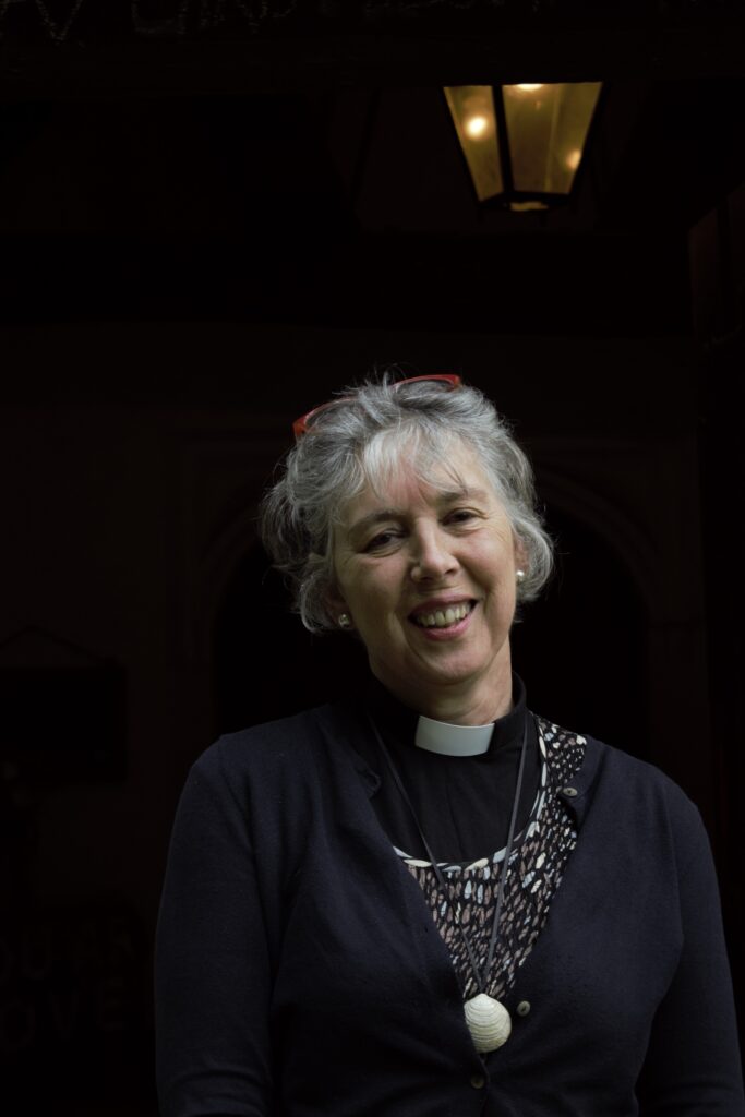 A portrait of a woman in black vlothes wearing a priest's collar. A Victorian-style lamp shines overhead.